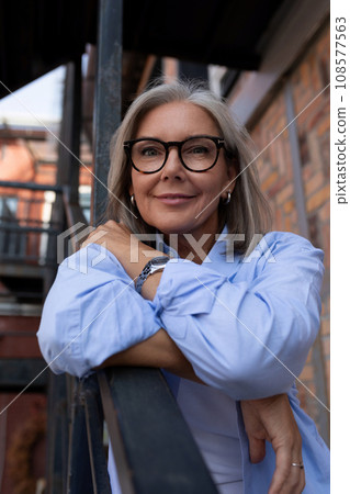 a mature woman with gray hair and glasses walks around the city in summer 108577563