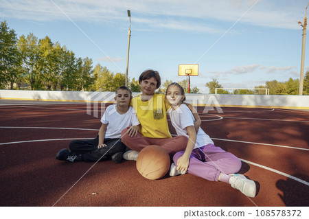 Outdoor hugs: Mother and teenage children cuddle gently in the sun on basketball court 108578372
