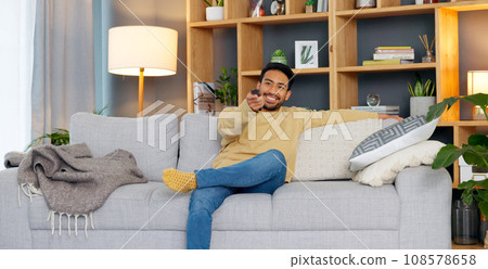 Watching tv, streaming and subscription with a man on a sofa in the living room of his home to relax. Television, video and remote with a young person changing the movie channel in his apartment Watching tv, streaming and subscription with a man on a sofa in the living room of his home to relax. Television, video and remote with a young person changing the movie channel in his apartment 108578658