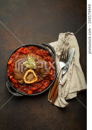 Traditional Italian dish Ossobuco all Milanese made with cut veal shank meat with vegetable tomato sauce served in black casserole pan top view on rustic brown background 108579949