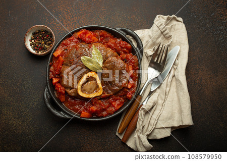Traditional Italian dish Ossobuco all Milanese made with cut veal shank meat with vegetable tomato sauce served in black casserole pan top view on rustic brown background 108579950