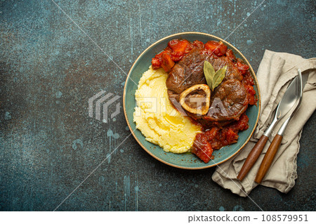 Traditional Italian dish Ossobuco all Milanese made with cut veal shank meat with vegetable tomato sauce served with corn polenta on ceramic plate top view on rustic stone background, copy space 108579951