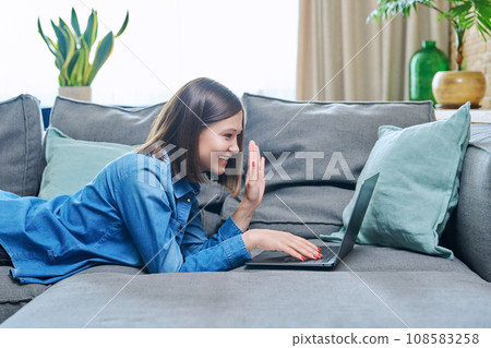 Young woman having video call lying on sofa at home, using laptop 108583258