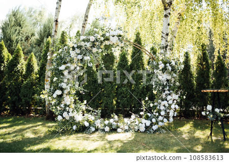 Circle wedding arch decorated with white flowers and greenery outdoors. 108583613
