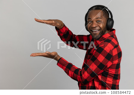 Cheerful black bearded man in headphones wearing red checkered shirt showing advertisement area Cheerful black bearded man in headphones wearing red checkered shirt showing advertisement area 108584472
