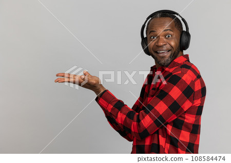 Amazed black bearded man in headphones wearing red checkered shirt presenting copy space on palm 108584474