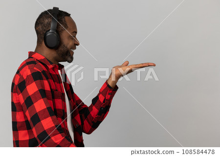 Smiling black bearded man in headphones wearing red checkered shirt presenting copy space on palm 108584478