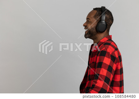 Profile of happy black bearded man in headphones wearing red checkered shirt listening to music Profile of happy black bearded man in headphones wearing red checkered shirt listening to music 108584480