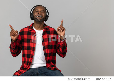 Smiling black bearded man in headphones wearing red checkered shirt pointing fingers up Smiling black bearded man in headphones wearing red checkered shirt pointing fingers up 108584486