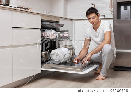 handsome man putting detergent tablet into dishwasher in kitchen handsome man putting detergent tablet into dishwasher in kitchen 108586728