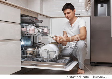 handsome positive man pulls out clean dishes from the dishwasher in white modern kitchen 108586729