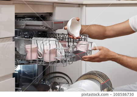 male hand unloading plates from open automatic dishwasher machine with clean utensils inside in kitchen. male hand unloading plates from open automatic dishwasher machine with clean utensils inside in kitchen. 108586730