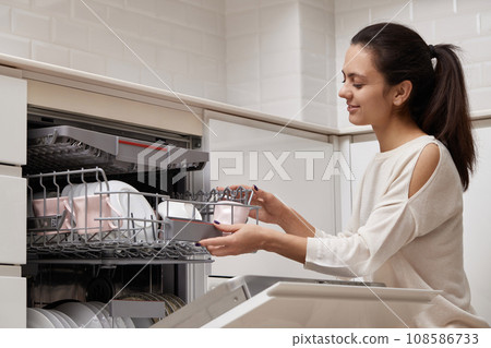 attractive woman unloading cup from open automatic built-in dishwasher machine with clean utensils inside in modern white kitchen. attractive woman unloading cup from open automatic built-in dishwasher machine with clean utensils inside in modern white kitchen. 108586733