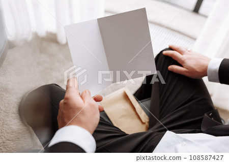 the groom holds an envelope with a letter from the bride. man is reading and sitting in the chair near the window. A letter for the groom. declaration of love. mock up place the groom holds an envelope with a letter from the bride. man is reading and sitting in the chair near the window. A letter for the groom. declaration of love. mock up place 108587427