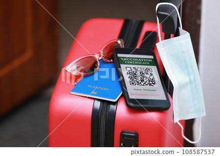 New normal concept. Close up view of a smartphone with an immune digital health passport, a red suitcase, protective mask, passport and sunglasses. Traveling by plane during a pandemic. 108587537
