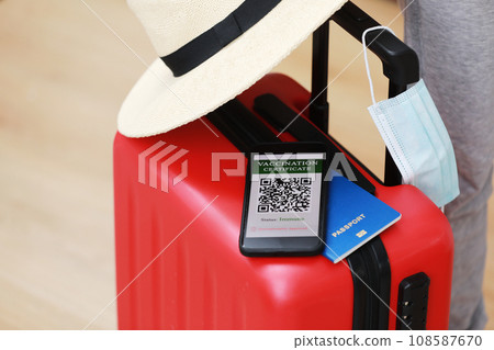 New normal concept. Close up view of a smartphone with an immune digital health passport, a red suitcase, protective mask, straw hat, passport and sunglasses. Traveling by plane during a pandemic. New normal concept. Close up view of a smartphone with an immune digital health passport, a red suitcase, protective mask, straw hat, passport and sunglasses. Traveling by plane during a pandemic. 108587670