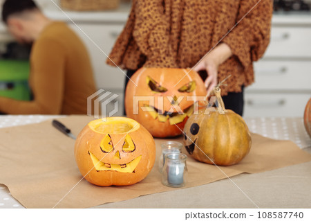 Close up of young woman carving Jack O Lattern from ripe orange pumpkin with knife on her wooden kitchen table. Female preparing all hallows eve Halloween party decorations. Background, copy space. Close up of young woman carving Jack O Lattern from ripe orange pumpkin with knife on her wooden kitchen table. Female preparing all hallows eve Halloween party decorations. Background, copy space. 108587740