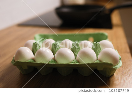 White chicken eggs in green cardboard egg tray on tabletop at modern stylish kitchen. Black frying wok pan on induction stove. Delicious healthy simple breakfast meal. Natural egg protein to the diet 108587749