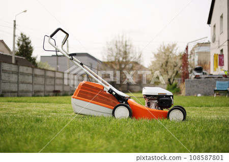 Modern orange-grey electric lawn mower on bright lush green lawn at residential backyard of house. Gardening work tools. Rotary lawn mower machine cut grass. Professional lawn care service. Modern orange-grey electric lawn mower on bright lush green lawn at residential backyard of house. Gardening work tools. Rotary lawn mower machine cut grass. Professional lawn care service. 108587801