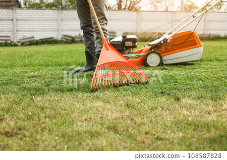 Male gardener collects cut grass with orange plastic rake after a mower, works in the backyard of the house. Man takes care of lawn. Concept of housework, gardening and country life, garden tools. Male gardener collects cut grass with orange plastic rake after a mower, works in the backyard of the house. Man takes care of lawn. Concept of housework, gardening and country life, garden tools. 108587828