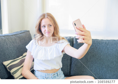 Caucasian girl sitting on the sofa and taking a selfie Caucasian girl sitting on the sofa and taking a selfie 108589277