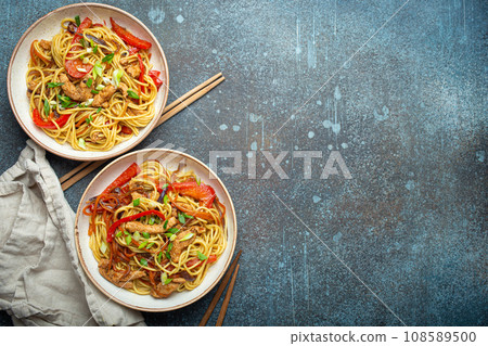 Two bowls with Chow Mein or Lo Mein, traditional Chinese stir fry noodles with meat and vegetables, served with chopsticks top view on rustic blue concrete background, space for text 108589500