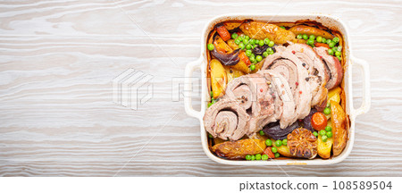 Rolled sliced pork roasted in white casserole dish with potatoes, vegetables and herbs on rustic white wooden background top view. Baked pork roll with vegetables for dinner 108589504