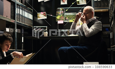 Senior agent working with criminologist, inspector feeling stressed about failing to find suspect. Partners analyzing new evidence and surveillance photos for culprit identification. Handheld shot. Senior agent working with criminologist, inspector feeling stressed about failing to find suspect. Partners analyzing new evidence and surveillance photos for culprit identification. Handheld shot. 108590633