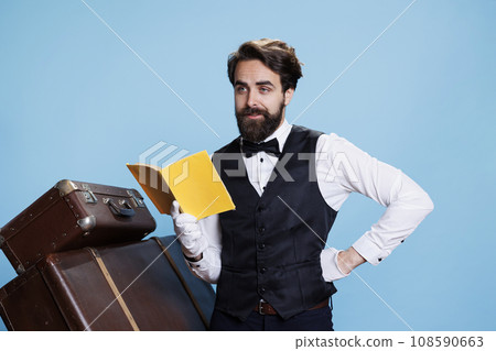 Hotel staff reads fiction book on camera, wearing tie and gloves while he is enjoying literature against blue background. Employee having fun with lecture hobby in studio, novel story. Hotel staff reads fiction book on camera, wearing tie and gloves while he is enjoying literature against blue background. Employee having fun with lecture hobby in studio, novel story. 108590663