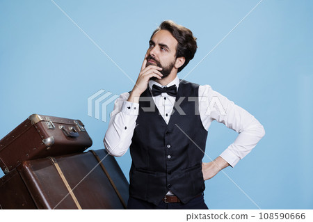 Hotel porter acts reflective in studio, thinking about something while he works in hospitality industry. Doorkeeper with tie posing next to luggage, being contemplative on camera. Hotel porter acts reflective in studio, thinking about something while he works in hospitality industry. Doorkeeper with tie posing next to luggage, being contemplative on camera. 108590666