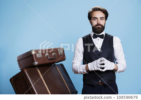 Classic bellhop with gloves and tie posing on camera, preparing to carry suitcases and give assistance to hotel guests. Young man working as doorkeeper or porter, hospitality industry. Classic bellhop with gloves and tie posing on camera, preparing to carry suitcases and give assistance to hotel guests. Young man working as doorkeeper or porter, hospitality industry. 108590679