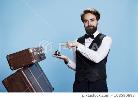Male bellhop with suitcases and bell, using to ring for service or assistance at hotel in hospitality industry. Young man working as doorman or bellboy handling trolley bags luggage. Male bellhop with suitcases and bell, using to ring for service or assistance at hotel in hospitality industry. Young man working as doorman or bellboy handling trolley bags luggage. 108590692