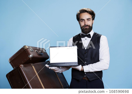 Hotel employee shows white screen on camera, holding laptop with blank copyspace display in studio. Professional luxury bellhop with tie presents chromakey template on pc, doorman. Hotel employee shows white screen on camera, holding laptop with blank copyspace display in studio. Professional luxury bellhop with tie presents chromakey template on pc, doorman. 108590698