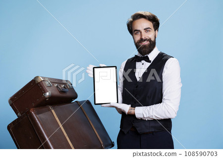 Bellboy presents white blank screen on tablet against blue background, wearing formal suit and gloves next to pile of suitcases. Classy porter showing copyspace template on device. Bellboy presents white blank screen on tablet against blue background, wearing formal suit and gloves next to pile of suitcases. Classy porter showing copyspace template on device. 108590703