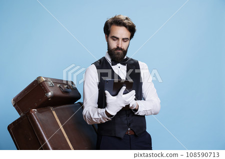 Bellboy with gloves reading reservations list on tablet, posing as a professional hotel porter next to trolley bags in studio. Doorkeeper in suit using device and arranging luggage for guests. 108590713