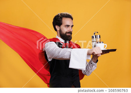 Superhero waiter carries food tray with red cape fluttering on camera, young man working at restaurant as butler serving dinner against yellow background. Professional elegant server. Superhero waiter carries food tray with red cape fluttering on camera, young man working at restaurant as butler serving dinner against yellow background. Professional elegant server. 108590763