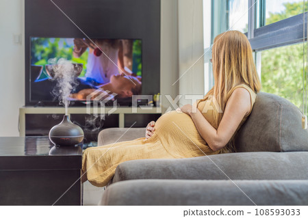 A blissful pregnant woman immerses in relaxation, savoring the soothing aroma from a diffuser while indulging in a calming TV video, embracing tranquility during her pregnant journey A blissful pregnant woman immerses in relaxation, savoring the soothing aroma from a diffuser while indulging in a calming TV video, embracing tranquility during her pregnant journey 108593033