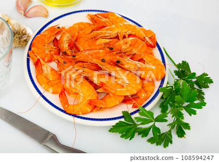 Plate with raw red shrimps, greens, condiments and knife on kitchen table Plate with raw red shrimps, greens, condiments and knife on kitchen table 108594247