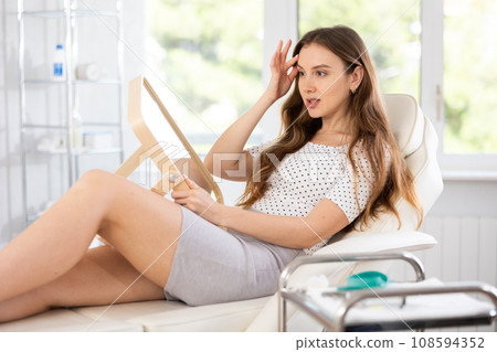 Satisfied young woman patient holding mirror lying on clinical chair Satisfied young woman patient holding mirror lying on clinical chair 108594352