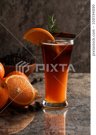 Iced Americano black coffee with lower layer orange juice in the glass. Rosemary with cinnamon on top. citrus fruits and coffee beans background. Coffee and orange juice concept. Iced Americano black coffee with lower layer orange juice in the glass. Rosemary with cinnamon on top. citrus fruits and coffee beans background. Coffee and orange juice concept. 108594890