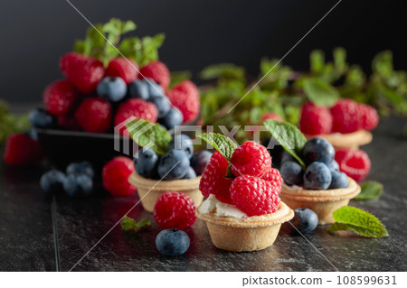 Small tartlets with fresh raspberries and blueberries on a black background. 108599631