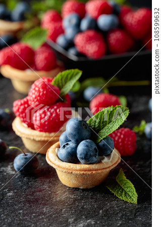 Small tartlets with fresh raspberries and blueberries on a black background. Small tartlets with fresh raspberries and blueberries on a black background. 108599632