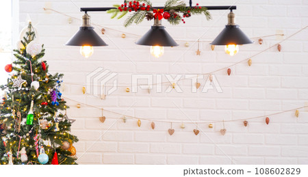 Christmas tree in white interior of a house with loft-style brick walls with garlands of glass toys on a rope. Glowing fairy lights decoration of the studio room. Cozy home in christmas and new year Christmas tree in white interior of a house with loft-style brick walls with garlands of glass toys on a rope. Glowing fairy lights decoration of the studio room. Cozy home in christmas and new year 108602829