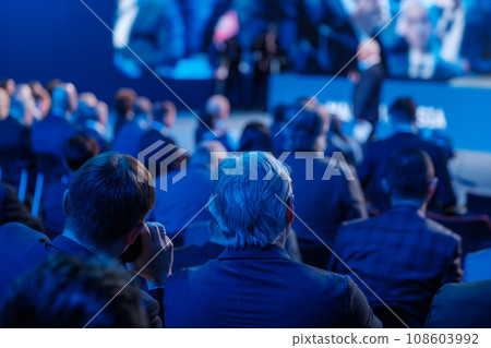 Businessmen listening to speaker giving presentation in illuminated auditorium at export forum  108603992