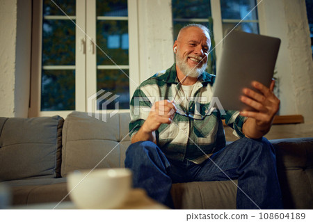 Portrait of mature man, grandfather sitting on coach in living room and surfing in Internet, shopping online in tablet and in headphones. Portrait of mature man, grandfather sitting on coach in living room and surfing in Internet, shopping online in tablet and in headphones. 108604189