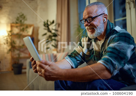 Side view portrait of mature man in glasses sitting on couch in living room and watching movie in tablet and in headphones on head. Side view portrait of mature man in glasses sitting on couch in living room and watching movie in tablet and in headphones on head. 108604190