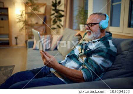 Side view portrait of senior man sitting on couch in living room and watching movie in tablet with headphones on head. Side view portrait of senior man sitting on couch in living room and watching movie in tablet with headphones on head. 108604193