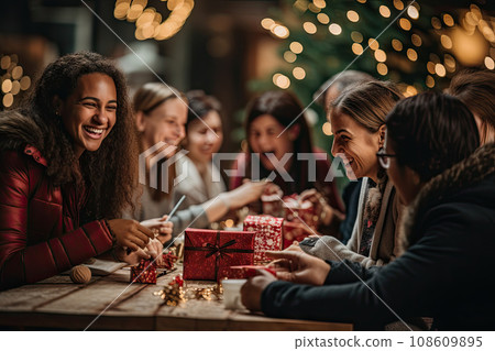 two women sitting at a table with christmas lights in the background and one woman holding a red box that says happy new year two women sitting at a table with christmas lights in the background and one woman holding a red box that says happy new year 108609895