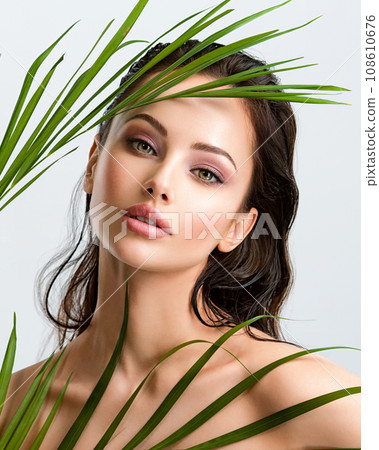 Young beautiful woman with healthy skin of face and palm leaves. Closeup fresh face of an attractive caucasian girl with green plants. Model with bright brown eye makeup. Skin care concept. Young beautiful woman with healthy skin of face and palm leaves. Closeup fresh face of an attractive caucasian girl with green plants. Model with bright brown eye makeup. Skin care concept. 108610676