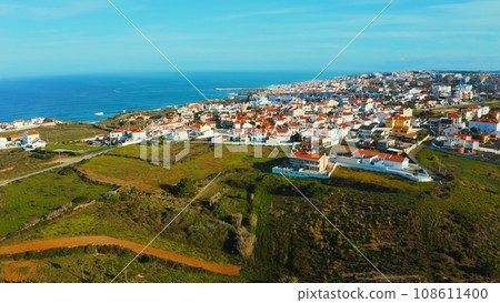 Aerial view of a small European town against blue sky and Atlantic Ocean. Drone view of a beautiful European city with a hilly landscape on ocean background. Beautiful natural landscape. Portugal. 108611400